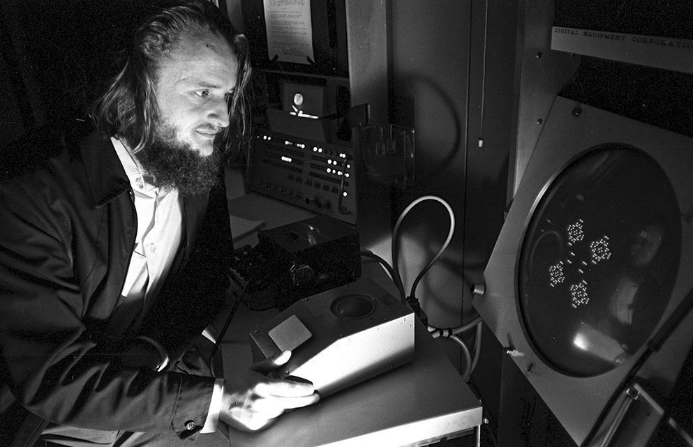 A black and white photo of mathematician John Conway watching a cellular automaton game that he invented, play out on a circular screen. 