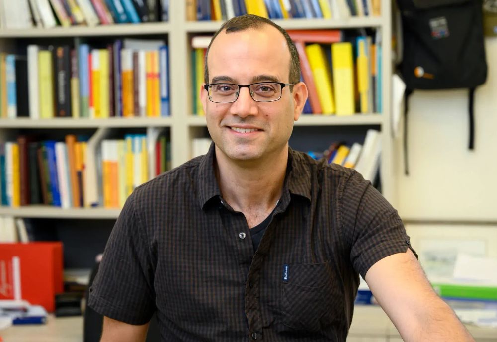 Mathematician Boaz Klartag smiles at the camera, sitting in front of a bookcase wearing a black plaid shirt 
