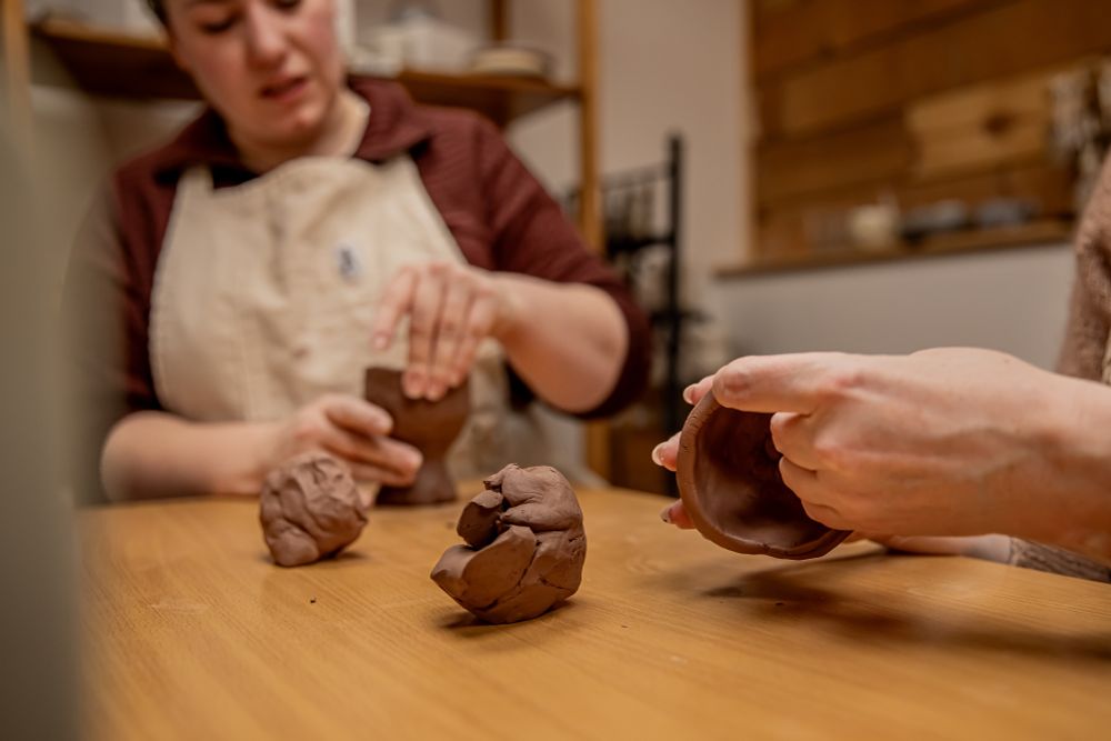 Two people sit at a table, squishing clay to make little pots in a ceramics studio.