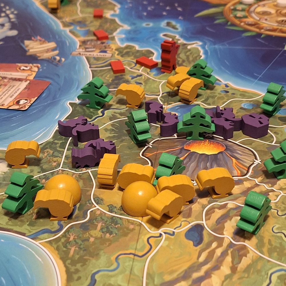 Kauri während der Partie. Spielsteine aus Holz aus Holz auf einem Inselplan. Gelbe Kiwis, lila Possums, rote Engländer und grüne Bäume.