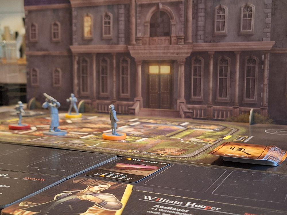 Terrorscape während der Partie von der Seite der Überlebenden aus gesehen. Im Hintergrund aufrecht eine große Villa aus Pappe. Davor ein Plan mit Räumen, auf dem verschiedene Miniaturen stehen. Eine davon hebt bedrohlich eine Kettensäge. Auf dem Plan liegt auch ein Stapel mit Spielkarten.  