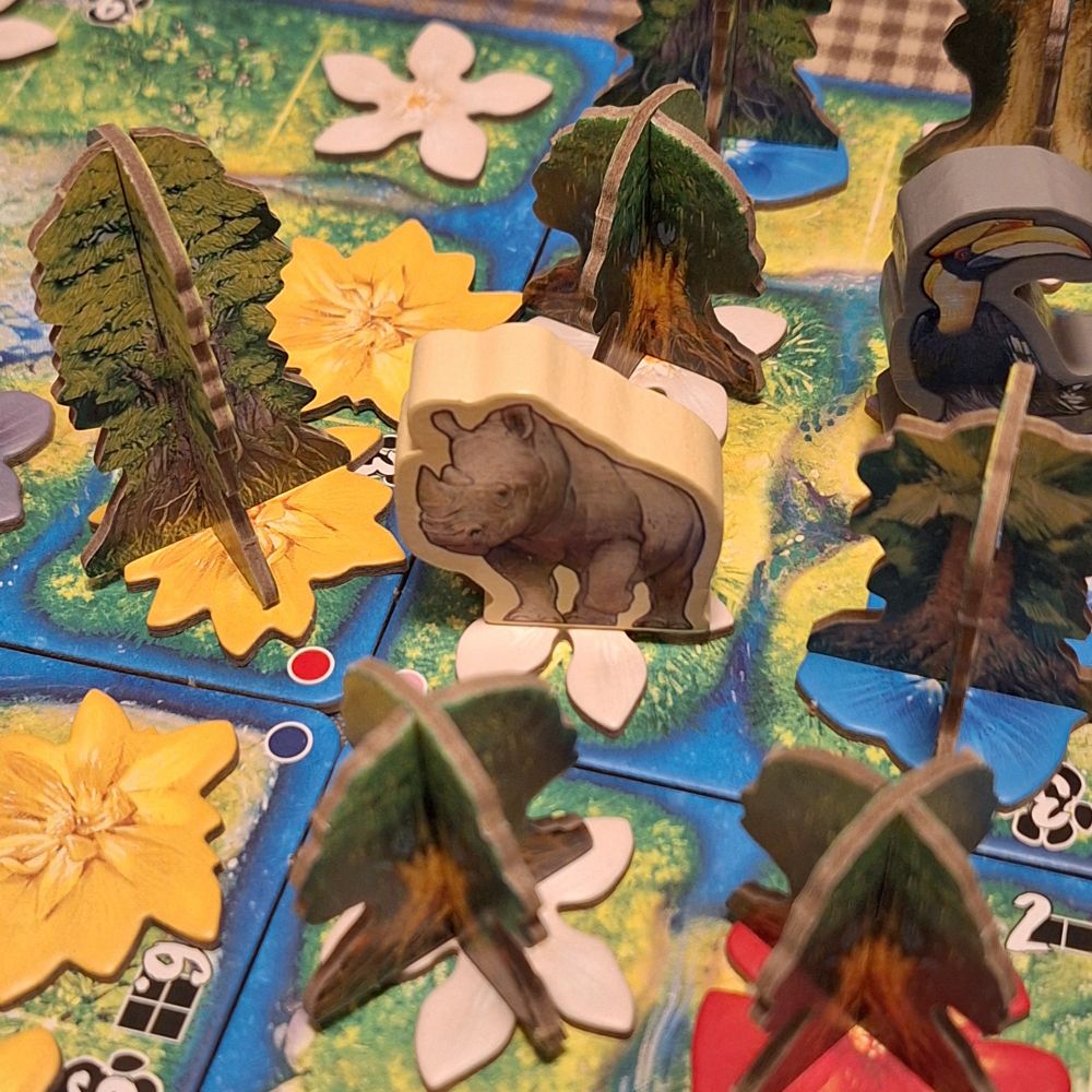 Hutan wahren das Spiels. In der Mitte ein bedruckter Nashorn-Token aus Holz, der auf einer weißen blute steht. Auf weiteren bunten Blüten darum stehen Pappaufsteller in der Form von Bäumen. 