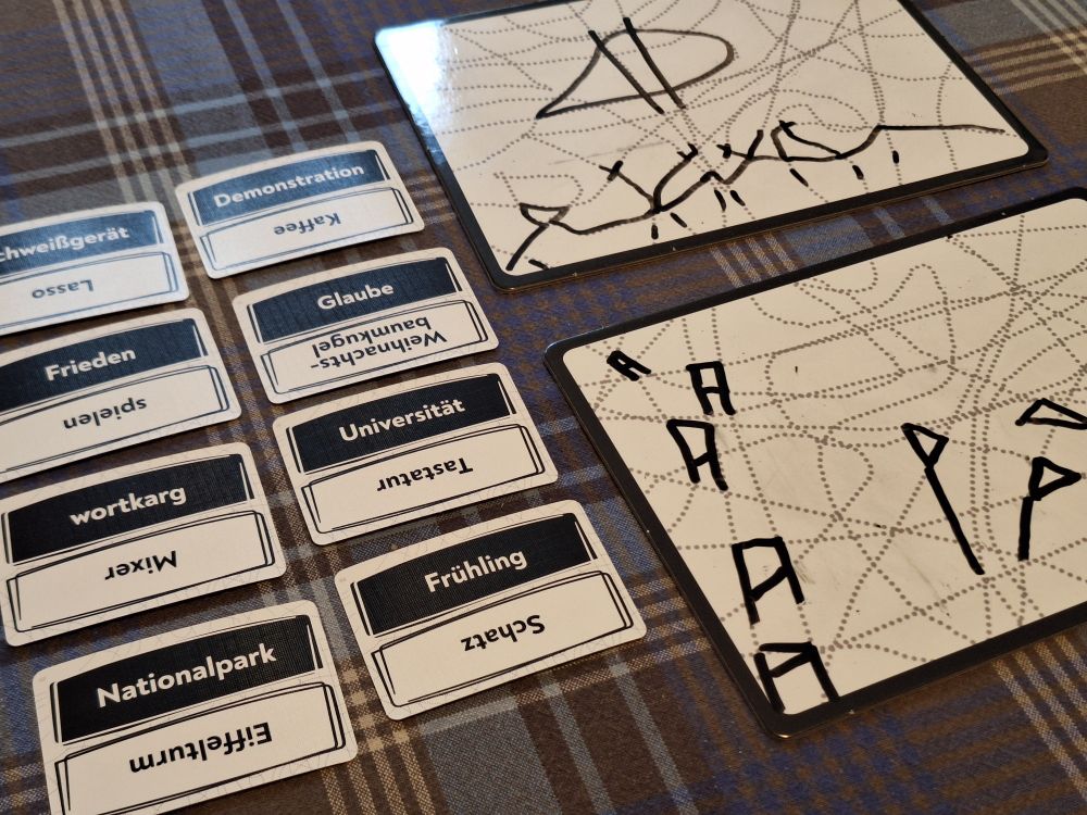 Krakel Orakel während der Partie: links Karten mit Worten darauf, rechts Pläne mit gestrichelten Linien, auf denen mit abwischbsrem Stift Begriffe gezeichnet wurden. Das Spiel ist komplett in Schwarz, Weiß und Grau gestaltet.