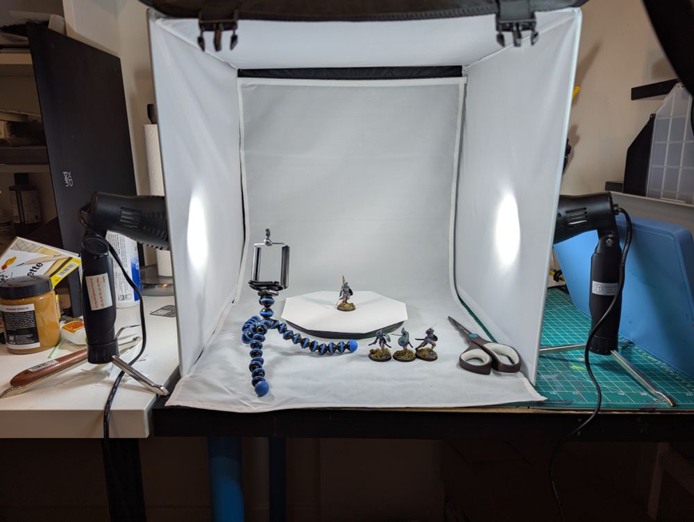 A portable photography both lit by portable lights from the left and right, occupying a messy hobby workstation. Inside the booth is a number of miniatures, a turntable, and a small gorilla pod tripod with a broken phone mount on top.