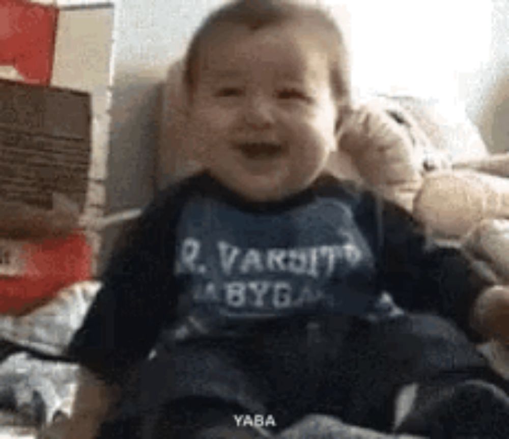 a baby is sitting on a bed wearing a varsity shirt and laughing .