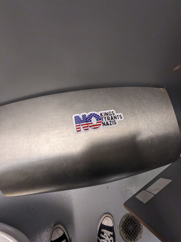 A sticker on a bathroom stall toilet paper dispenser that says "no kings, no tyrants, no Nazis"