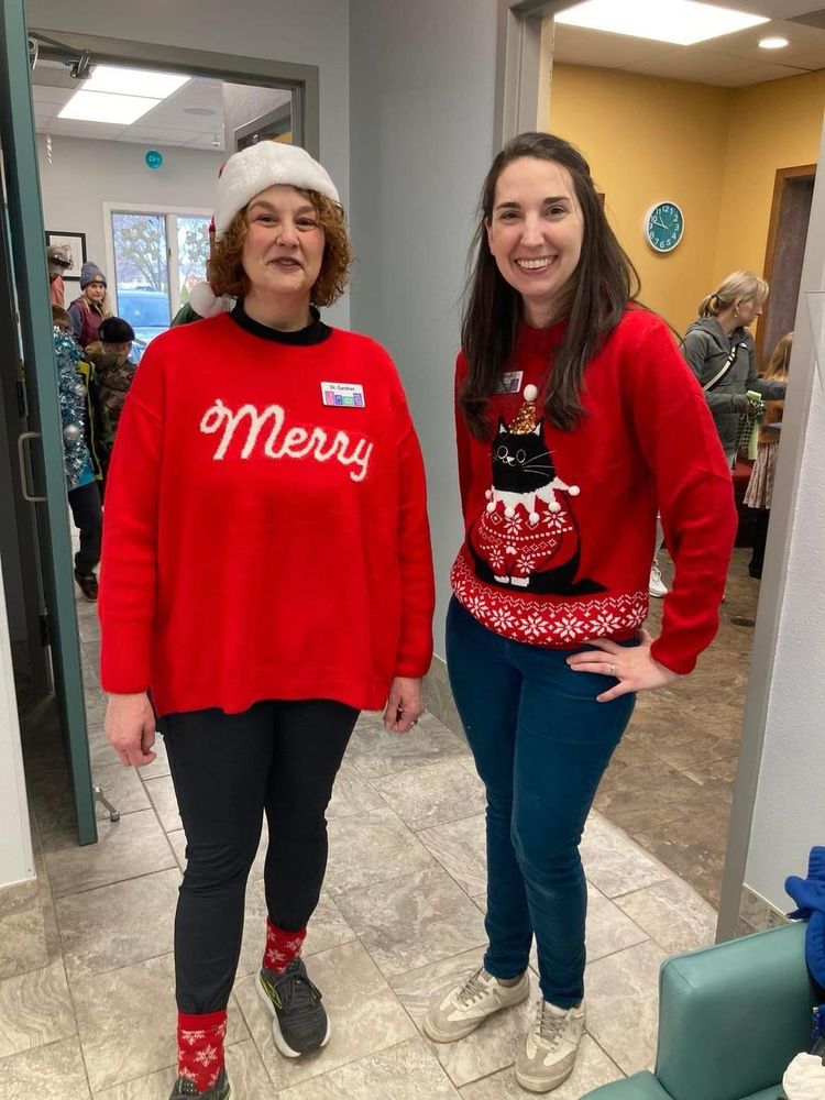 two women posing for picture, standing in the middle of a busy veterinary clinic, both wearing bright red holiday sweaters