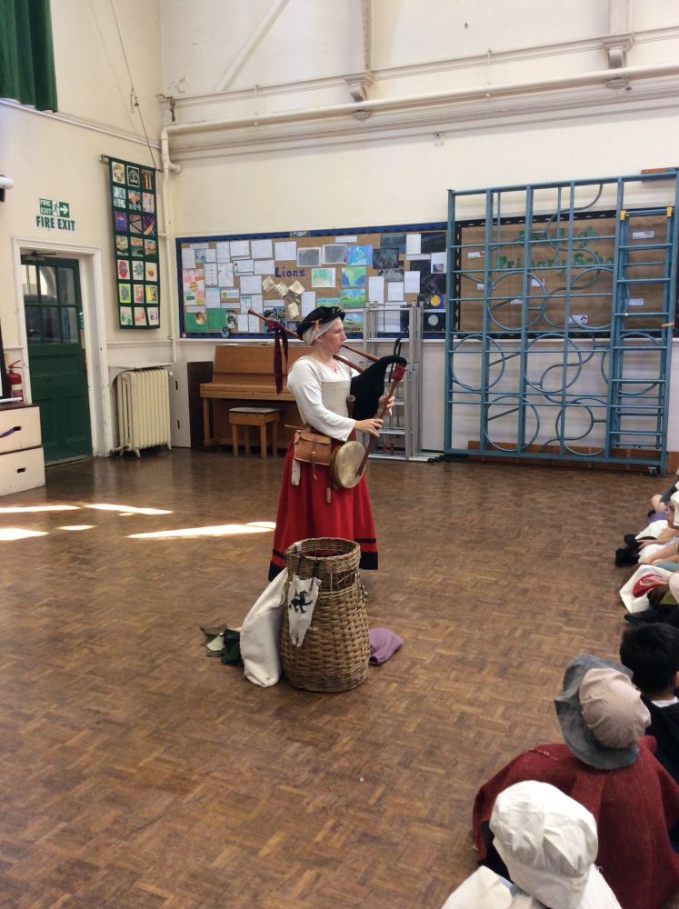 Emma is standing in a school hall, dressed in authentic Tudor clothing playing the bagpipes. Next to her is a basket with other instruments in, in front of her are seated primary school children. 