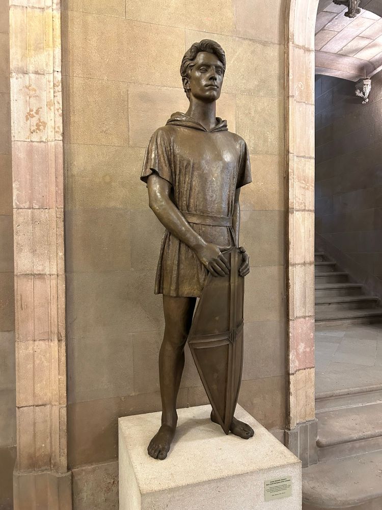 Bronze statue of someone that looks a bit like Peter Pan, in a robe holding a shield