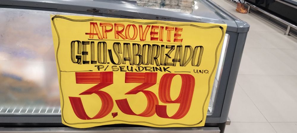 Cartaz de supermercado afixado ao lado de um freezer da seção de frios, feito de cartolina amarela fom letras manuscritas que indicam "Aproveite. Gelo saborizado. Para seu drink. Unidade. 3,39.