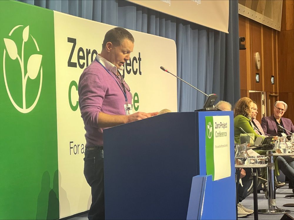 Side image of Jerry standing behind a podium.  Several panelists are in the background