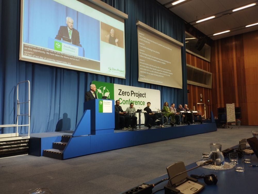View of the main conference stand.  A man is standing at the podium and is projected on a large screen.  7 panelists are seated to his right.