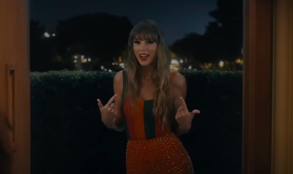 A screenshot of the antihero music video. Showgirl Taylor is standing outside the door in sparkly orange shorts and an orange and green striped top, pointing to herself as she sings “I’m the problem, it’s me”