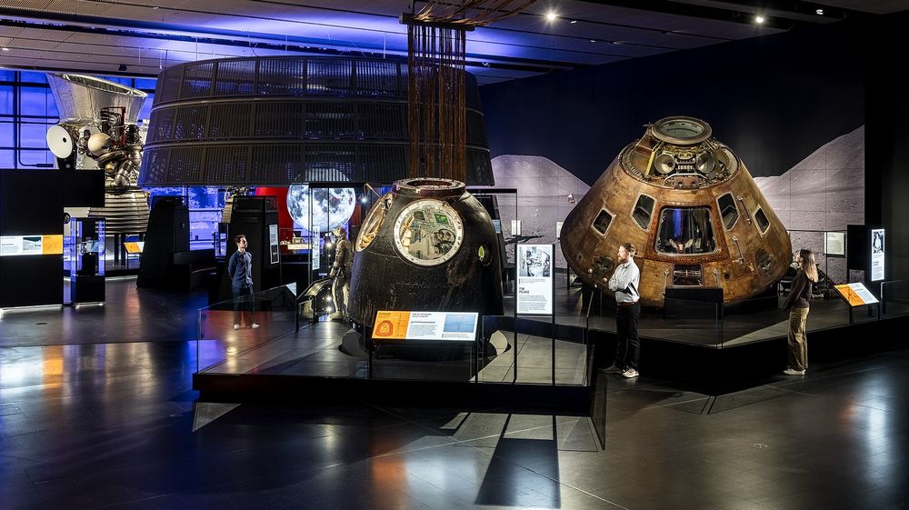 Visitors stand in the Science Museum's new Space gallery, observing an Apollo 10 command module, a Soyuz space capsule and a spacesuit. Also prominent are a 3D projection of the Moon and large rocket and satellite components. 
