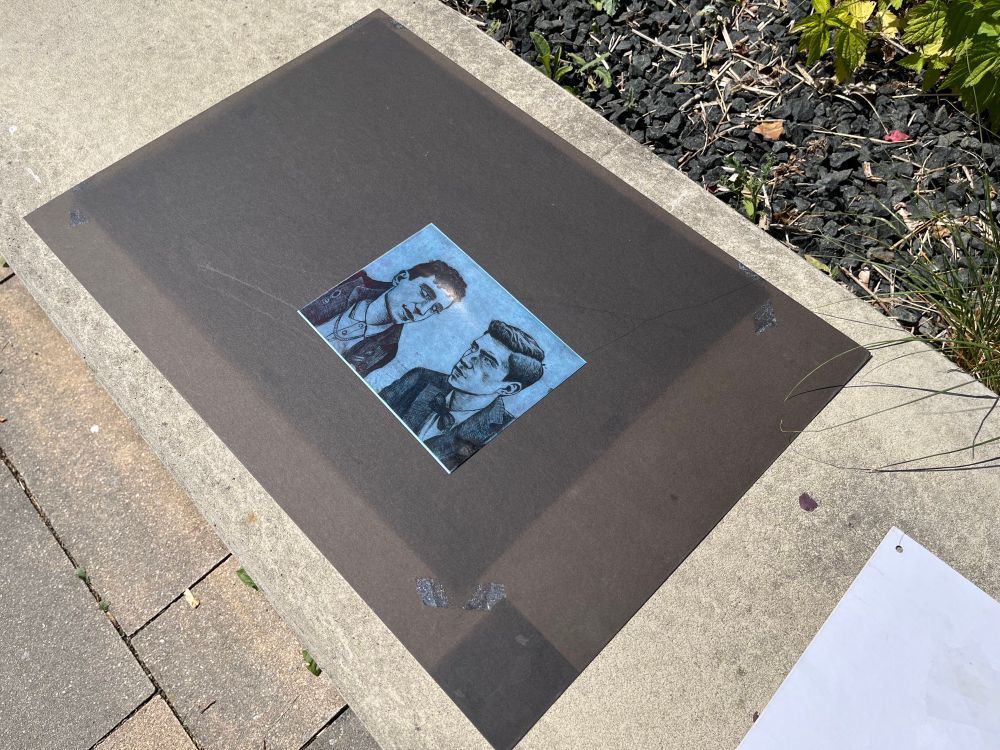 Photo of black cardboard lying in the sun. On it is blue cyanotype paper with a printed transparent etching plate showing Charles and Edwin looking at each other.