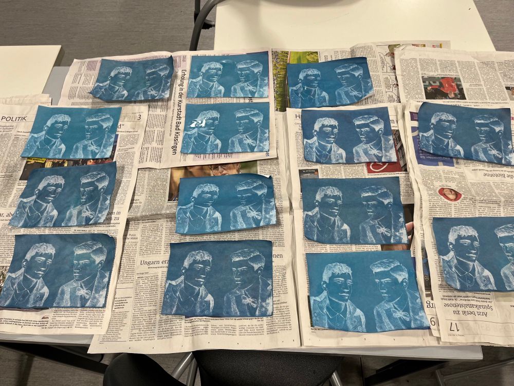 Photo of 14 sheets of cyanotype paper drying on newspapers after the photosensitive surface has been fixed with water. The motif is white now, while the cyanotypes have various shades of blue.