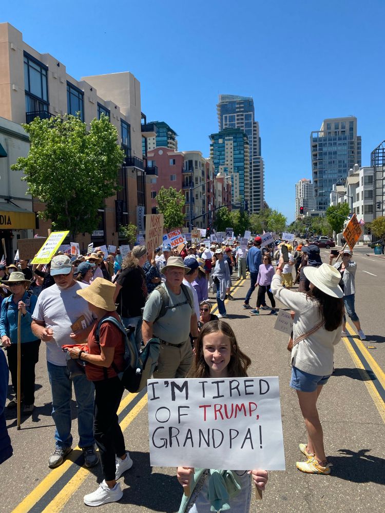 Young girl standing in front of a large protest group, holding a sign saying “I’m tired of Trump, Grandpa!”