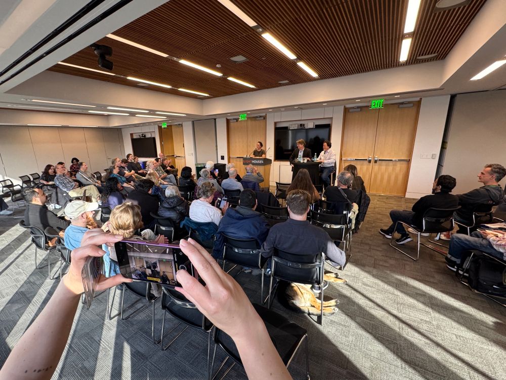 An audience sits in rows of black chairs, listening to Josh and Jason’s discussion, while someone in the foreground takes a photo.