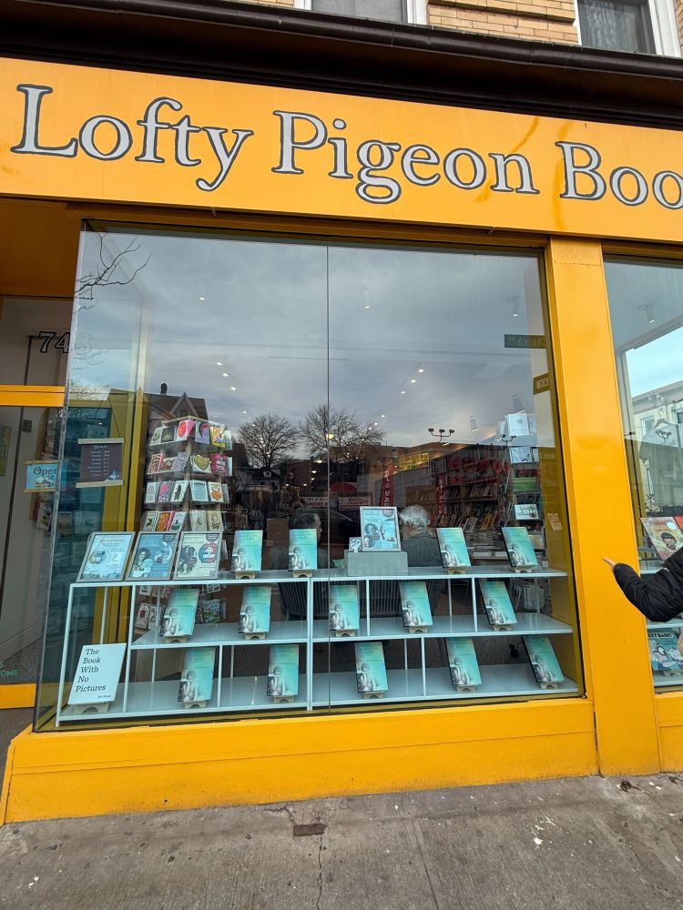 The exterior of Lofty Pigeon Books shows a yellow storefront with large glass windows. The window display features multiple copies of Connecting Dots - A Blind Life, arranged neatly on white shelves.