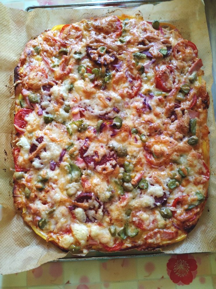 Домашняя пицца на столе на бумаге для запекания. Прямоугольная с закруглёнными углами. Содержит сырокопчёную колбасу, сыр, помидоры, оливки