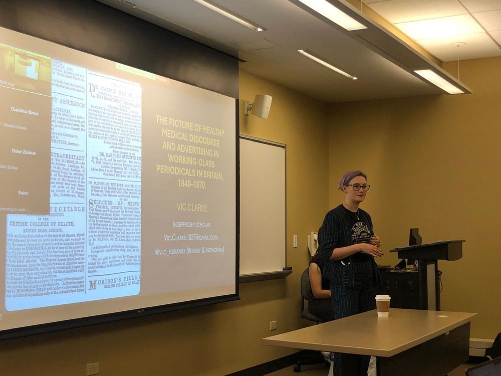 A picture of me (a woman with purple hair) presenting my paper, in front of a PowerPoint slide adorned with Victorian healthcare adverts.