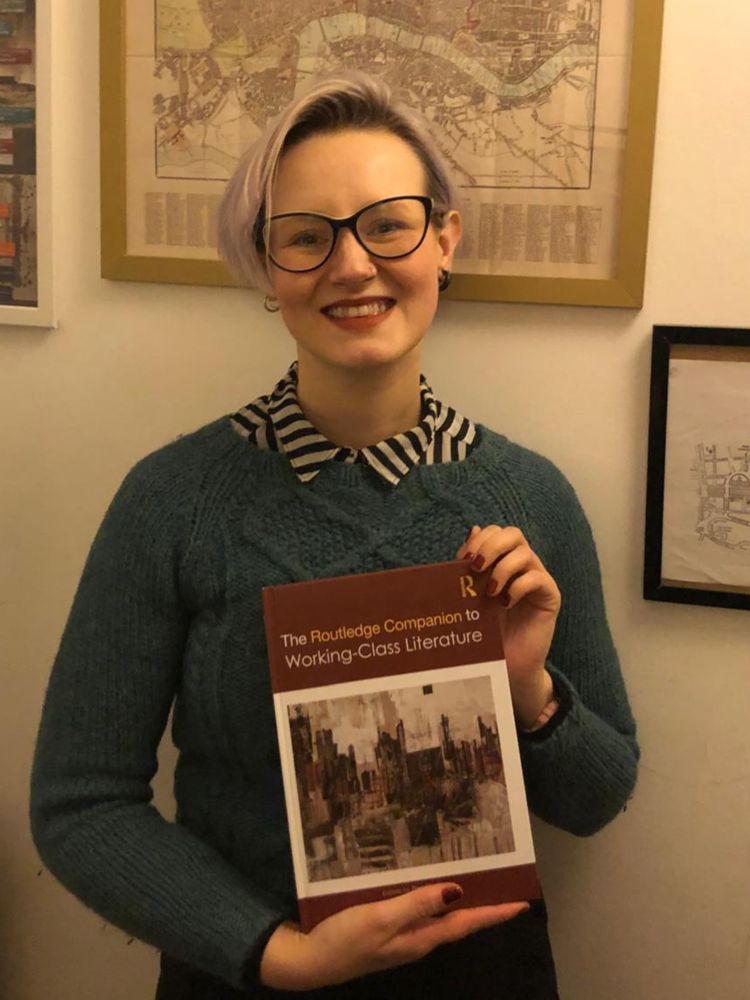 Vic is a woman with glasses and purple hair, smiling and holding her copy of the Routledge Companion to Working-Class Literature.