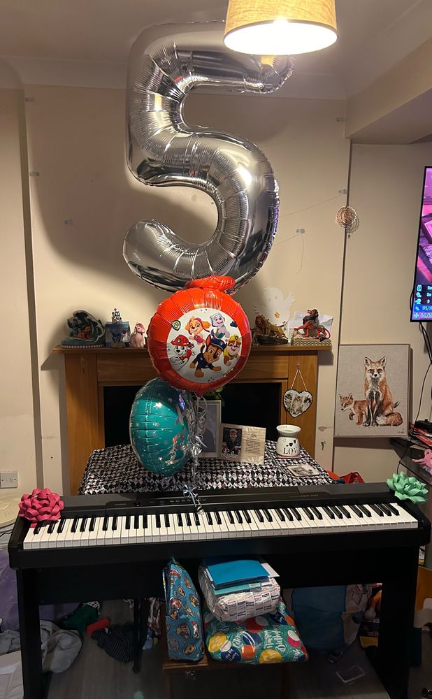 Foil balloons hover about an electric piano with presents on a table in front.