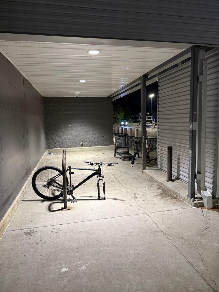 Bike rack in well lit area, hidden from main store entrance, with a lone, sad bike, locked up, missing its front wheel and saddle 