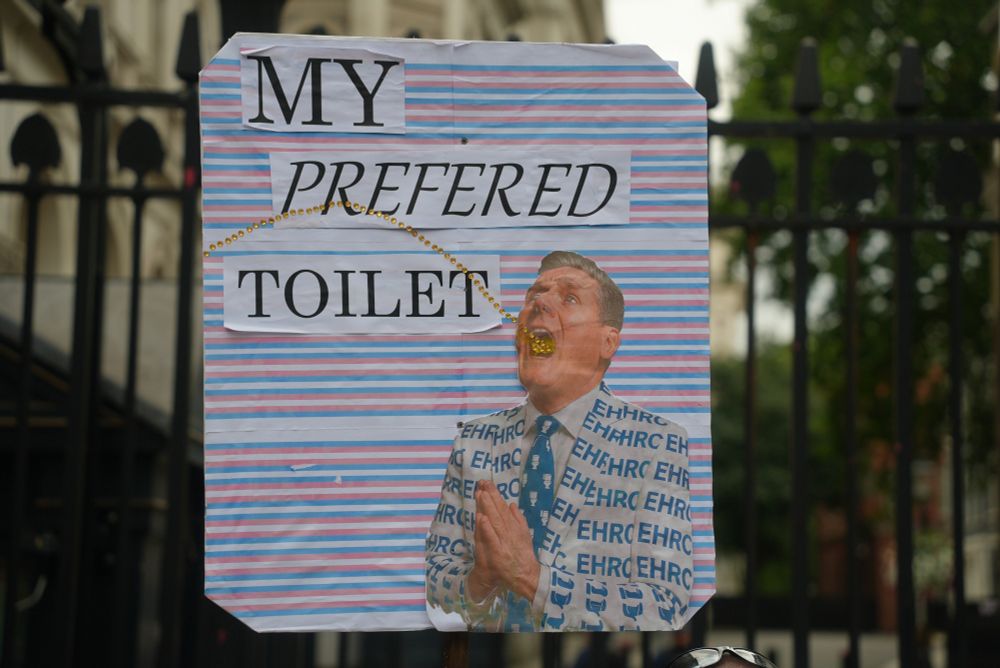 A protest sign saying "My preferred Toilet" over a picture of Keir Starmer with his mouth open, wearing an EHRC jacket, and a line of yellow glass beads leading into his mouth like droplets. The photo was taken outside the barrier to Downing Street at London Trans+ Pride 2025.