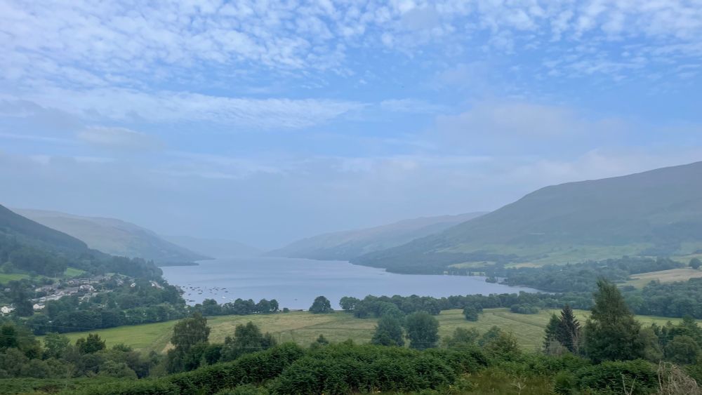 View down loch from high on hillside with blue sky but hazy hills and end of loch