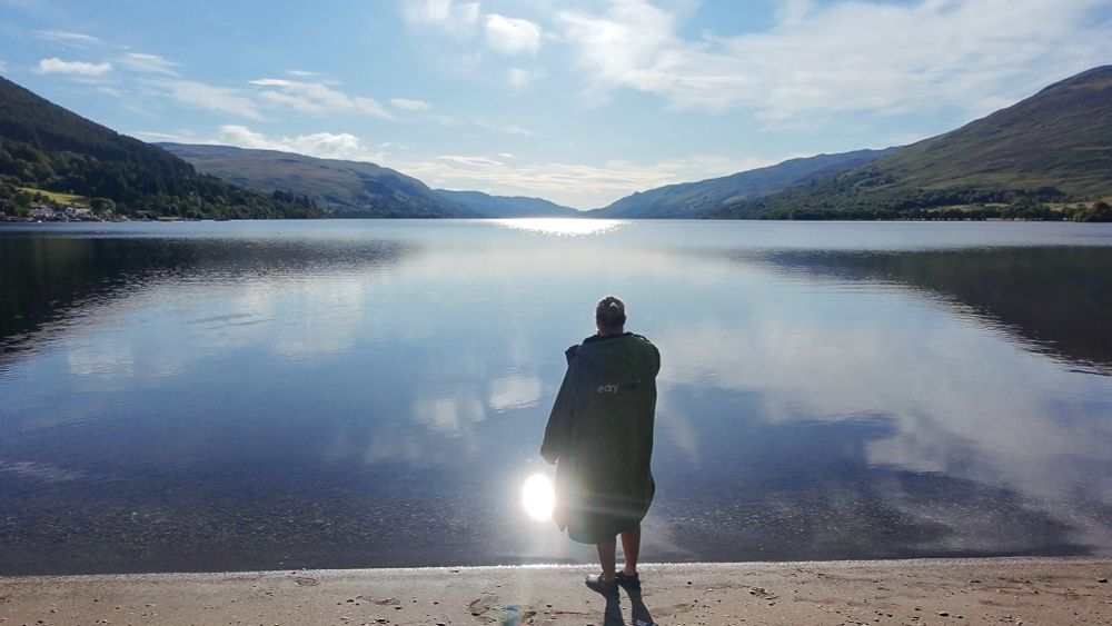 Me stood on shore in robe with blue water of loch and some clouds in sky. 