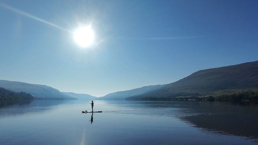Very still loch with no clouds and just sun and silhouette of a paddle boarder  