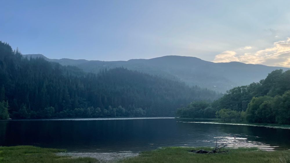 Blue sky and sun setting. Bottom third is dark still water, second third is forest and then sky above. 