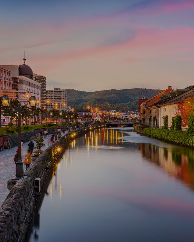 [Image description: Image shows an evening captured in Otaru, Hokkaido Japan, showcasing the canal with city streets on each side and pink sunset across the water.]