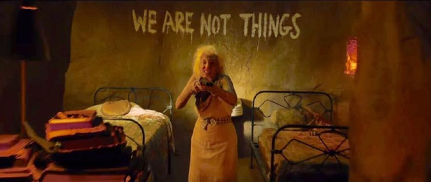a still from mad max fury road where an older woman is pointing a blunderbuss type gun at the camera. She is in a sparse bedroom with several small beds. Above her, on the wall, horror movie style letters read “WE ARE NOT THINGS”