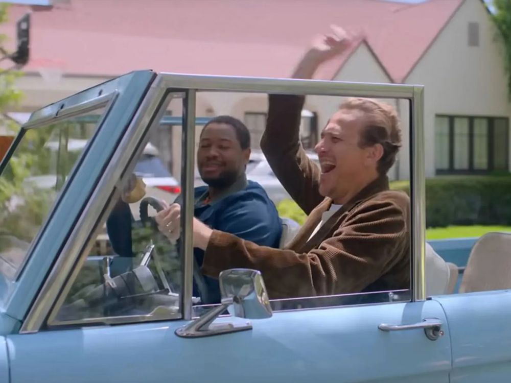 Still from film #Shrinking. Jimmy driving his Ford Bronco, raising his arm in the air in celebration, with Sean in the front seat smiling at him.