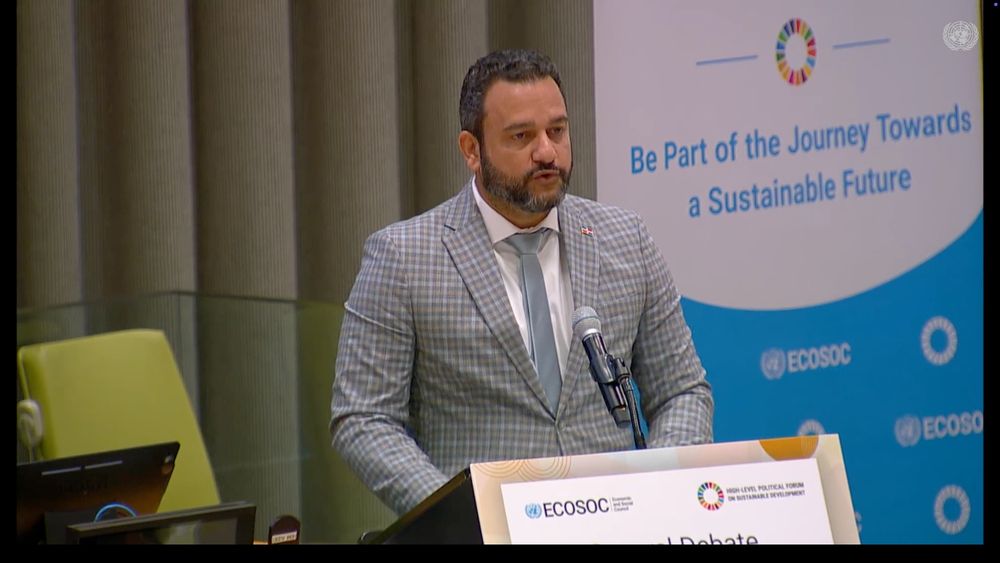 H.E. Luis Madera, VM for Monitoring and Government Coordination of the Ministry of the Presidency of the Dominican Republic speaking into a podium wearing a blue checkered suit and tie.
