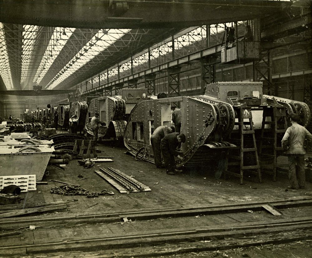 A black-and-white photograph of the interior of a tank factory. Tanks are lined up into the distance, with men working on them.