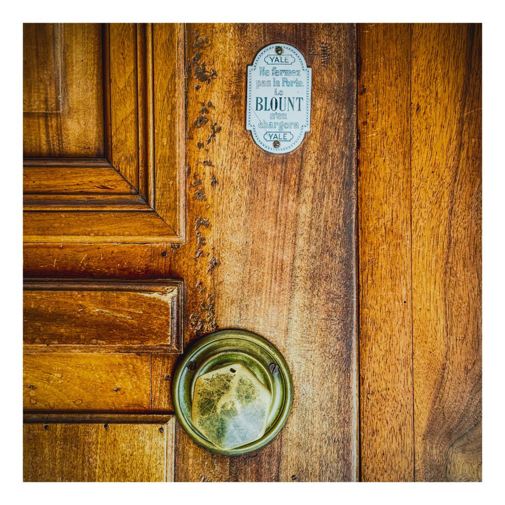 A tightly cropped frame of a worn hexagonal metal door handle on a varnished wood doo with a white ceramic sign telling people not to close the door.