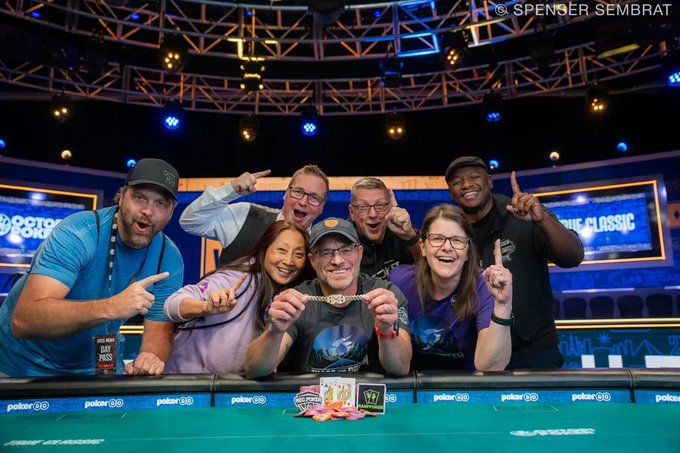 Craig's winner's photo. Craig is sitting at a poker table behing a pile of chips and the winning hand, a pair of Queens. He is smiling and holding up his bracelet. Six people are leaning in behind him, holding up one finger. Among these individuals are Carlos Welch and Dayanna Ciabaton.