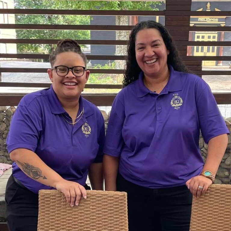 Michelle Farmer and Natascha Carpenter, both Black women in matching purple shirts, are standing behind a poker table, smiling.