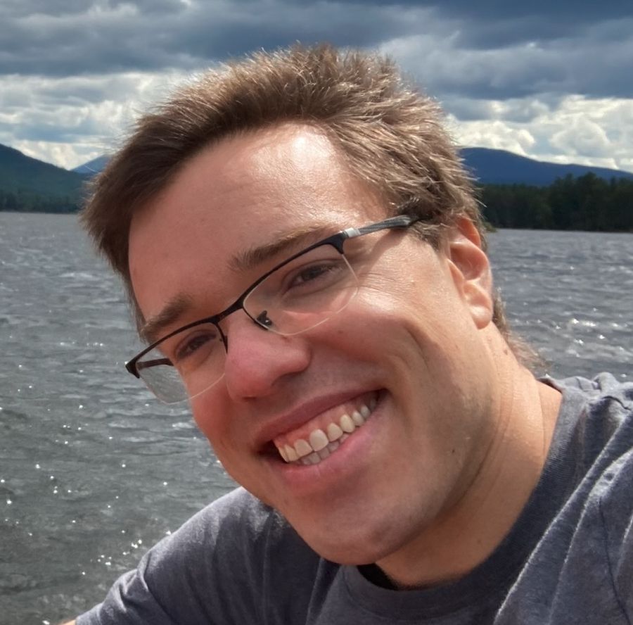 Nate Meyvis in front of a lake on a sunny day. Nate is a middle-aged white man with glasses, short brown hair, a strong jaw line, and a toothy smile. His head is tilted approximately 45 degrees to the right.