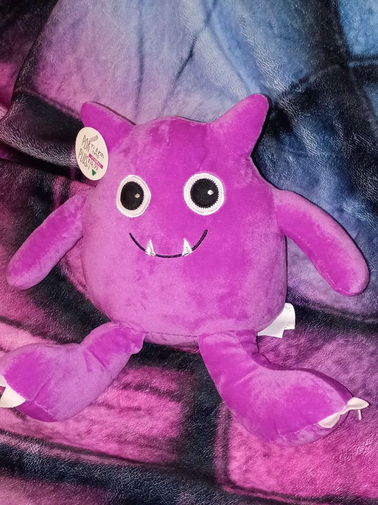 shot of a plushie on a blanket. The plushie is a pink adorable demon thing  with big eyes a smile, and two upward pointing fangs