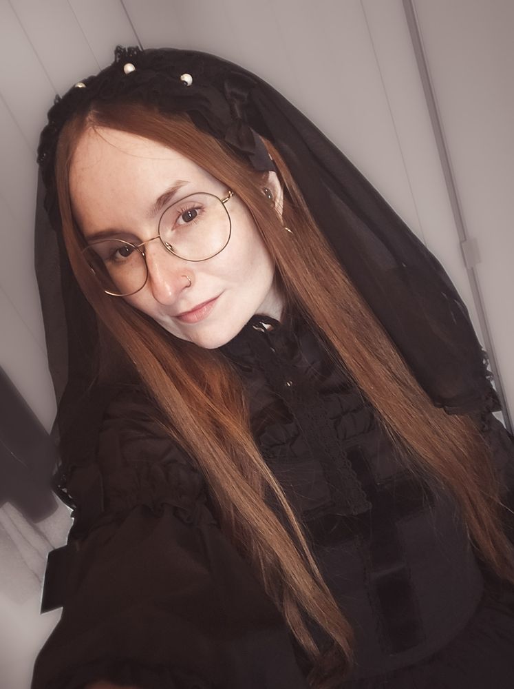 Selfie to showcase the black veiled headdress with pearls on top by Angelic Pretty. Long natural red hair is framing the face, and large round glasses are framing the eyes. The black velvet cross on the bodice of the jsk is also seen more clearly in this photo.
