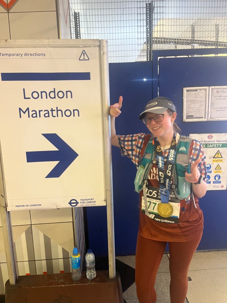 Alex posing with bisexual finger guns by a London Marathon sign post-race