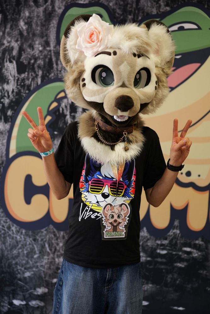A picture of me wearing my partial fursuit, which is a white and grey hyena named Fresnel, while standing in front of the chomp event banner.