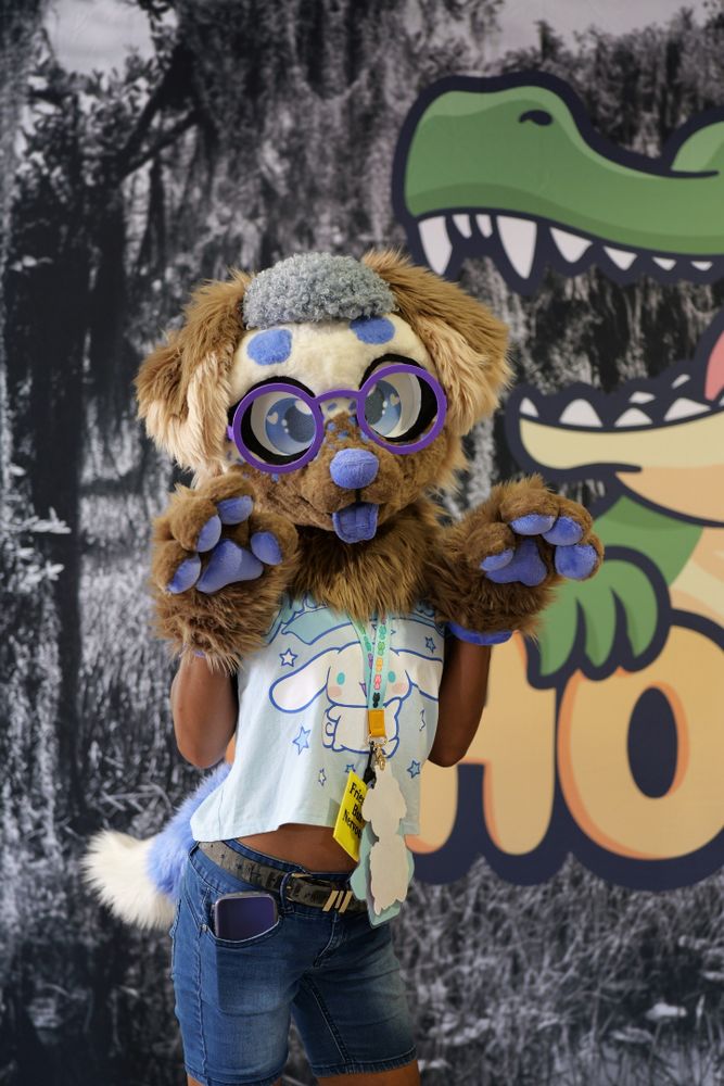 A brown canine fursuiter with purple glasses posing in front of the chomp event sign.