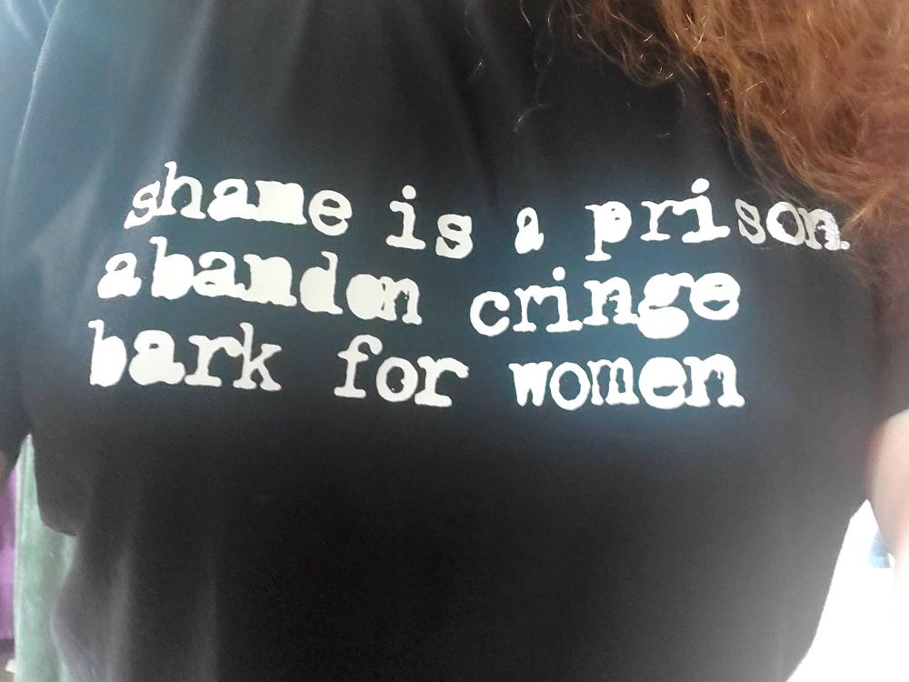a photograph of me (specifically my tits) wearing a black shirt that in white print says "shame is a prison. Abandon cringe. Bark for women" a bit of my hair is visible draped over my chest.