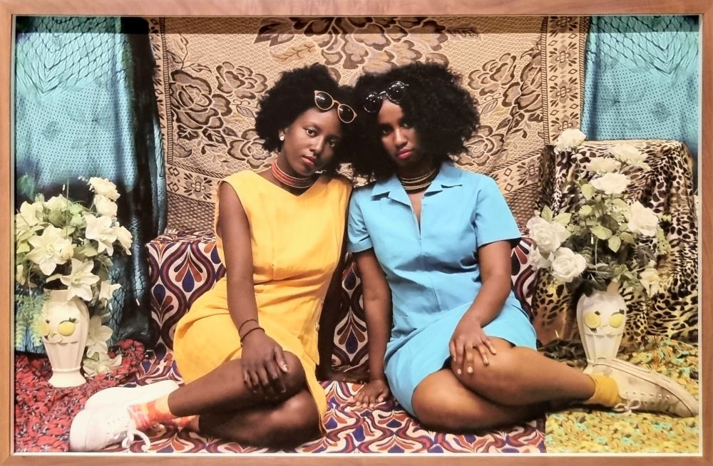 A photo of two black women. They are sitting on colourful patterned rugs, one dressed in blue and one in yellow, and they press their black hair together. They have sunglasses propped up on their heads. There are flowers in vases on either side.