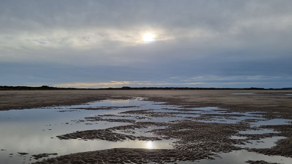 Weak sunshine reflected in tide pools on Holkham beach. A line of dunes is on the horizon.